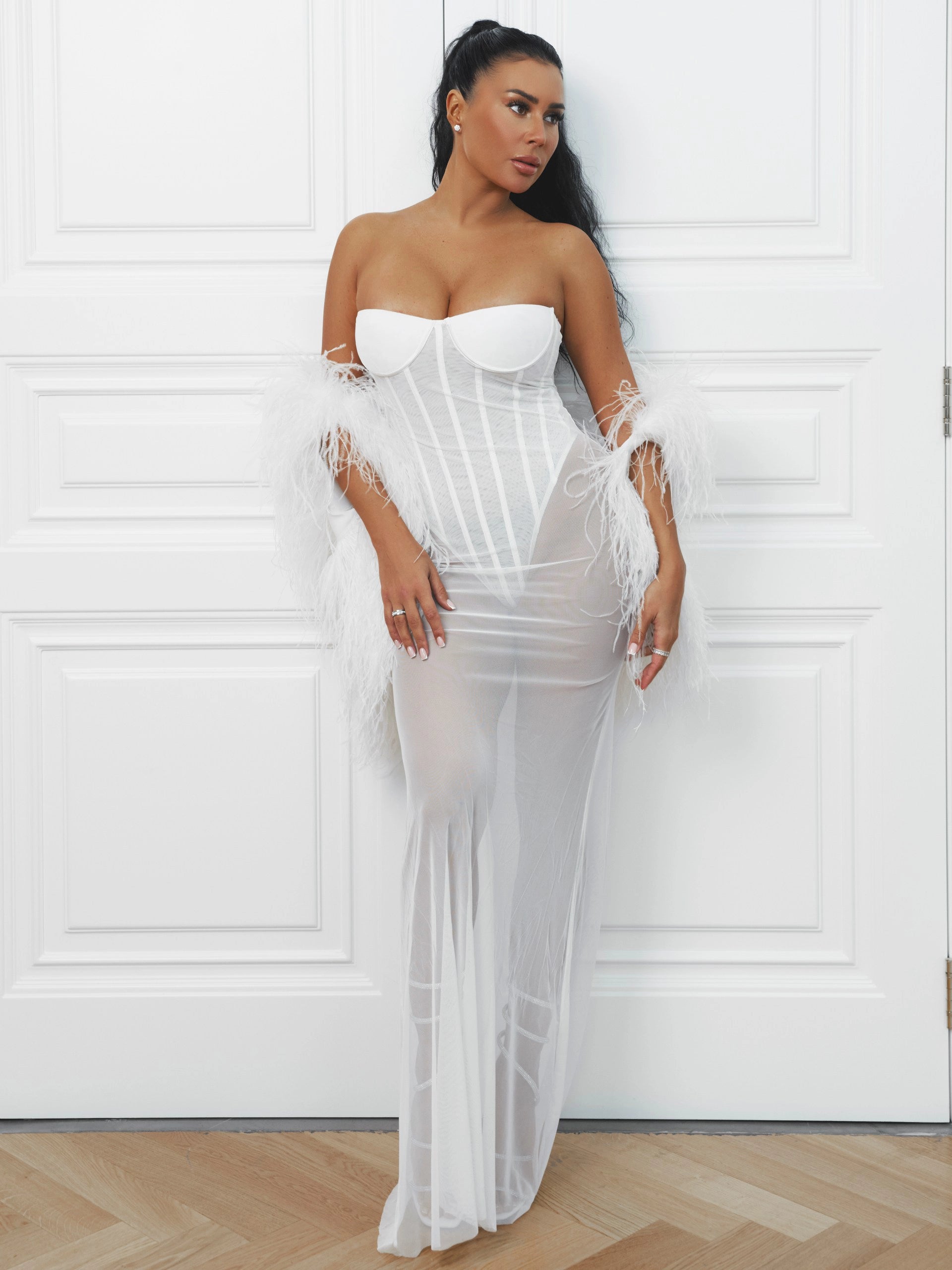 Woman wearing a white strapless gown with sheer overlay against a white wall.