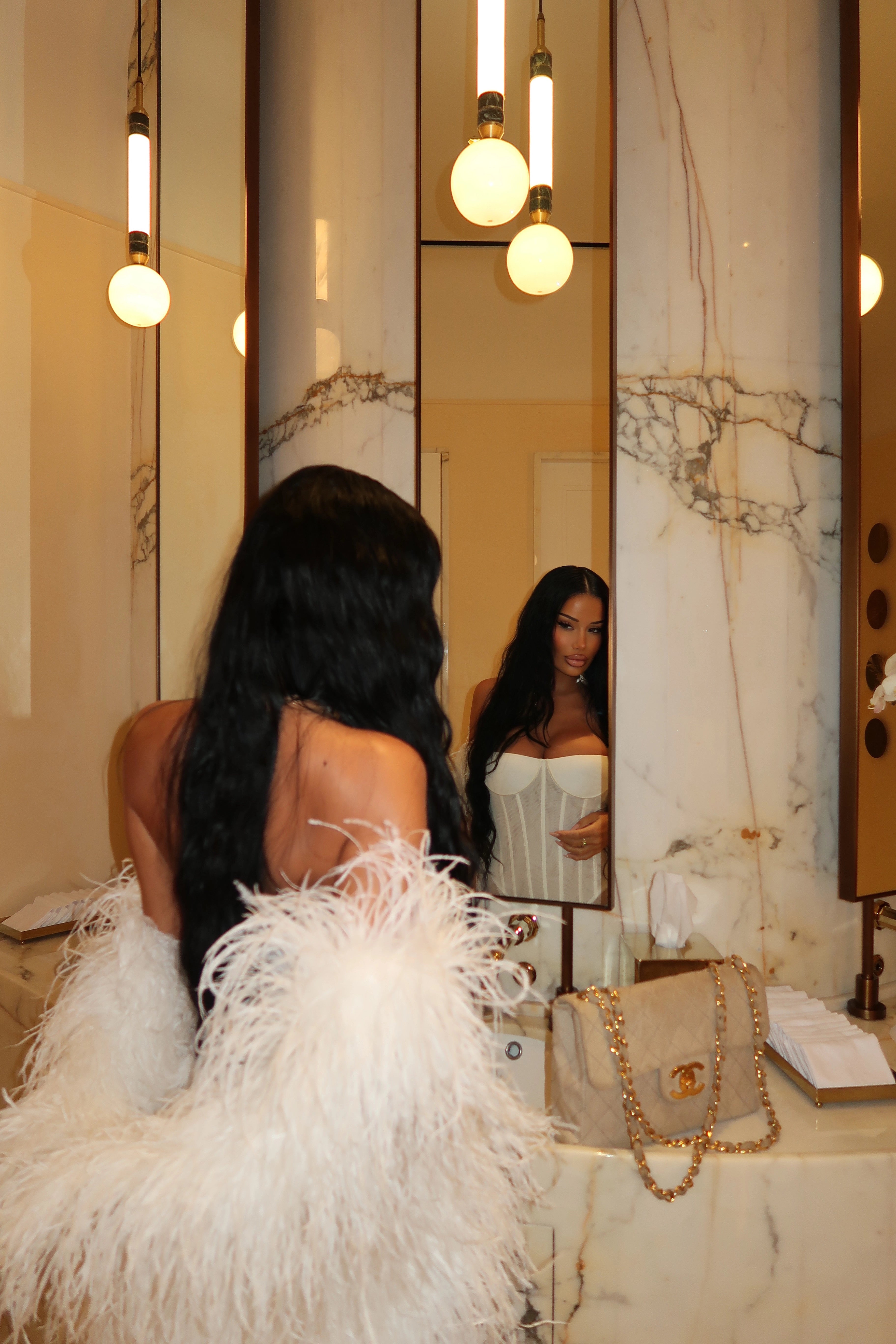 Woman in a white feathered dress looking at herself in a mirror with a gold handbag on a table.