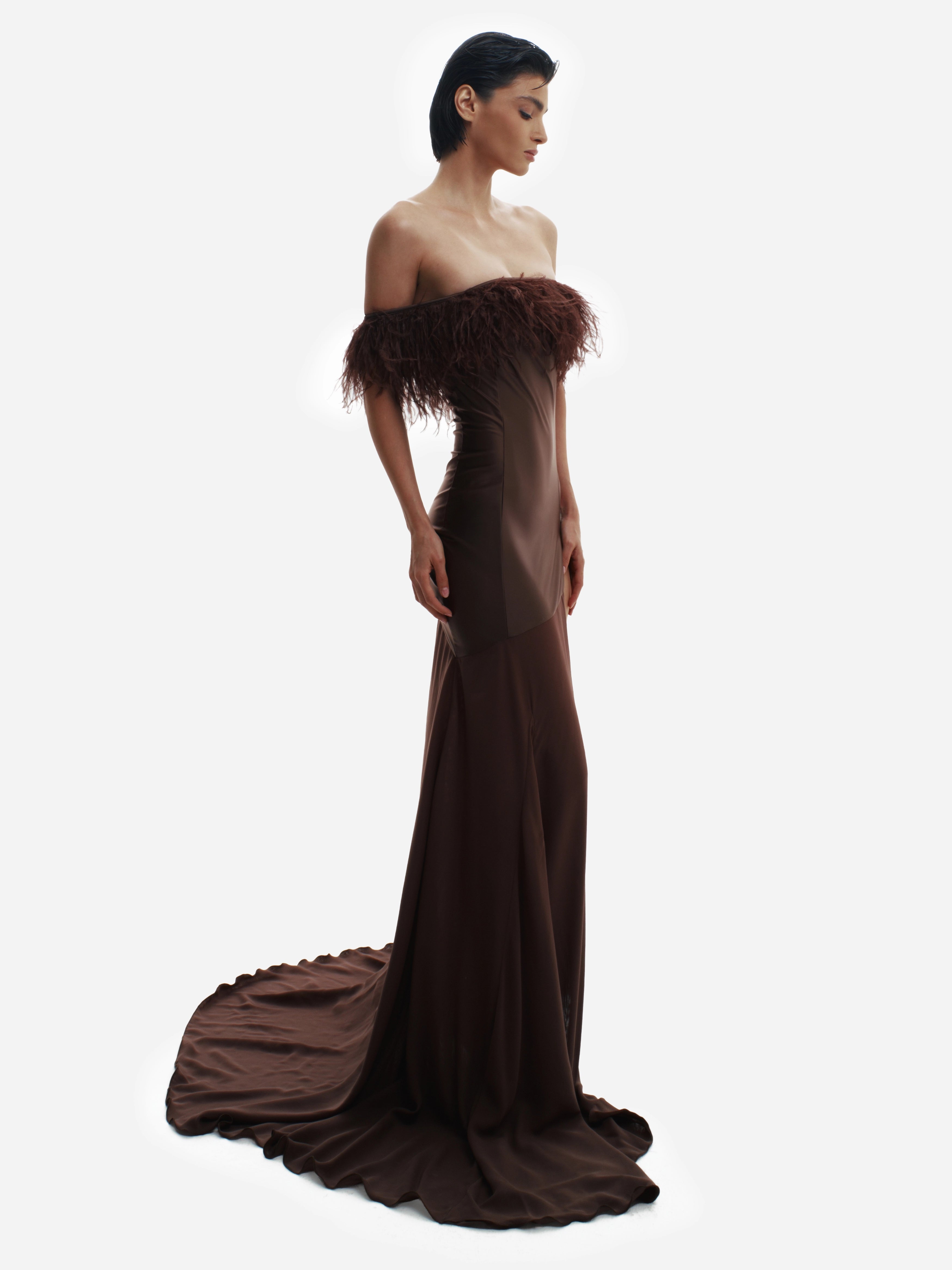 Woman wearing a dark brown off-shoulder gown with feathered details on a white background